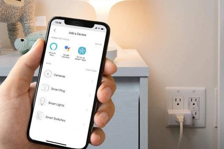 گوشی هوشمندی که در دست است، برنامه Kasa Smart Plug را نمایش می‌دهد، با دوشاخه برق یک چراغ رومیزی در پس‌زمینه.
